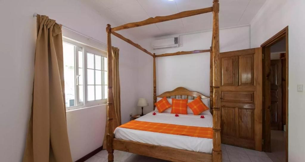 Una cama con dosel con una colcha blanca y naranja, un cabecero de madera y almohadas naranjas. Se observa una ventana con cortinas color beige y una puerta de madera al fondo.