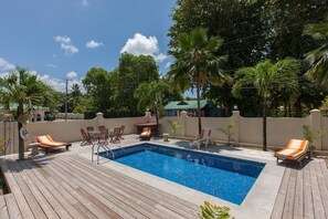A swimming pool with lounge chairs and a wooden deck, surrounded by palm trees and a clear sky. - Villa Confort (Praslin Island)