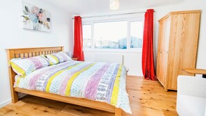 A bedroom with a wooden bed, a window with red curtains, a radiator, and a wooden wardrobe.