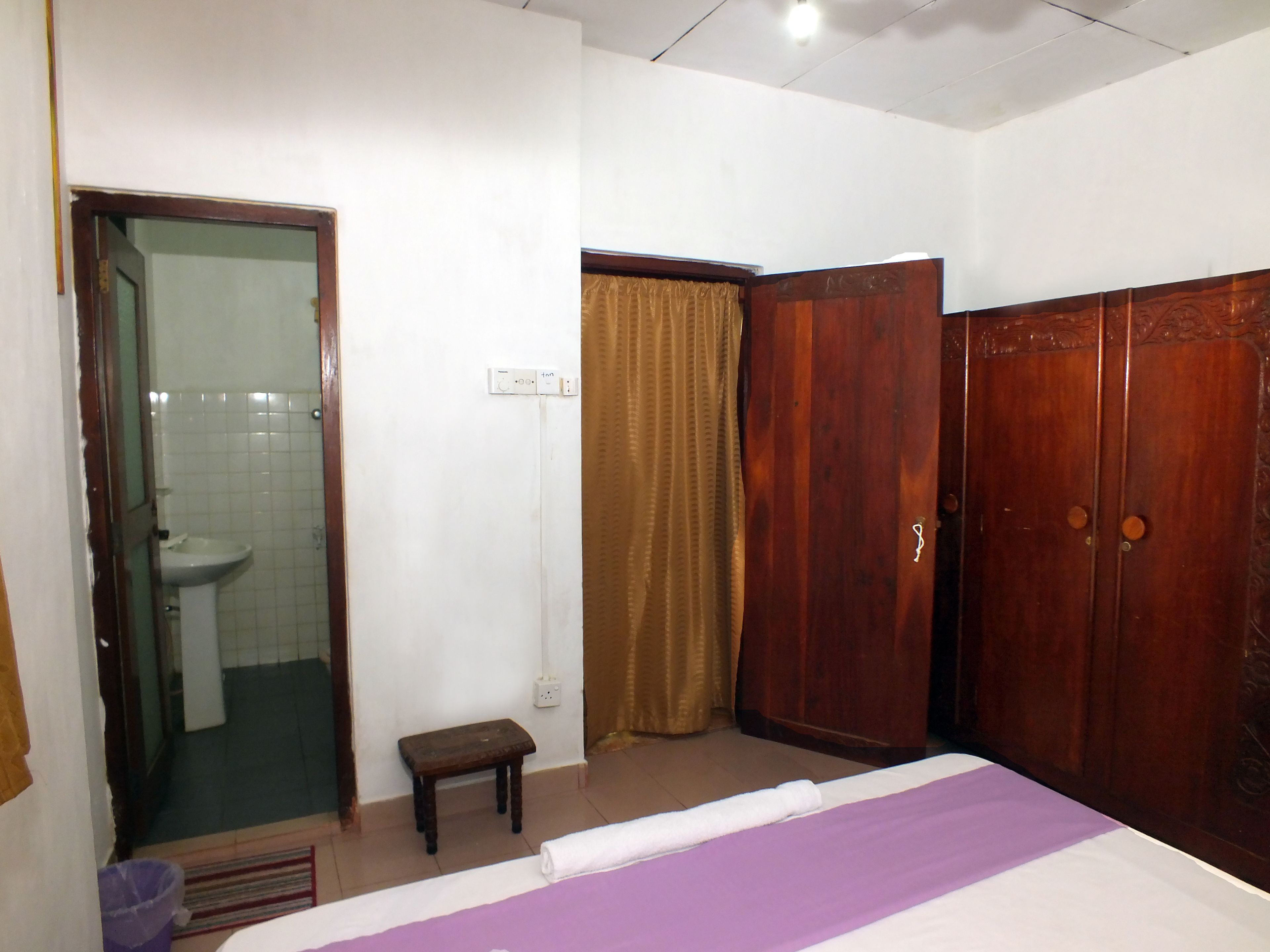 A bedroom with a bed, a wooden wardrobe, a bathroom with a toilet and sink, and a small wooden stool.