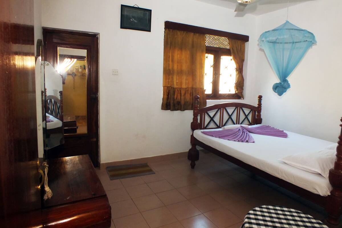 A bedroom with a bed, a wooden wardrobe, a ceiling fan, a window with curtains, and a small rug on the floor.