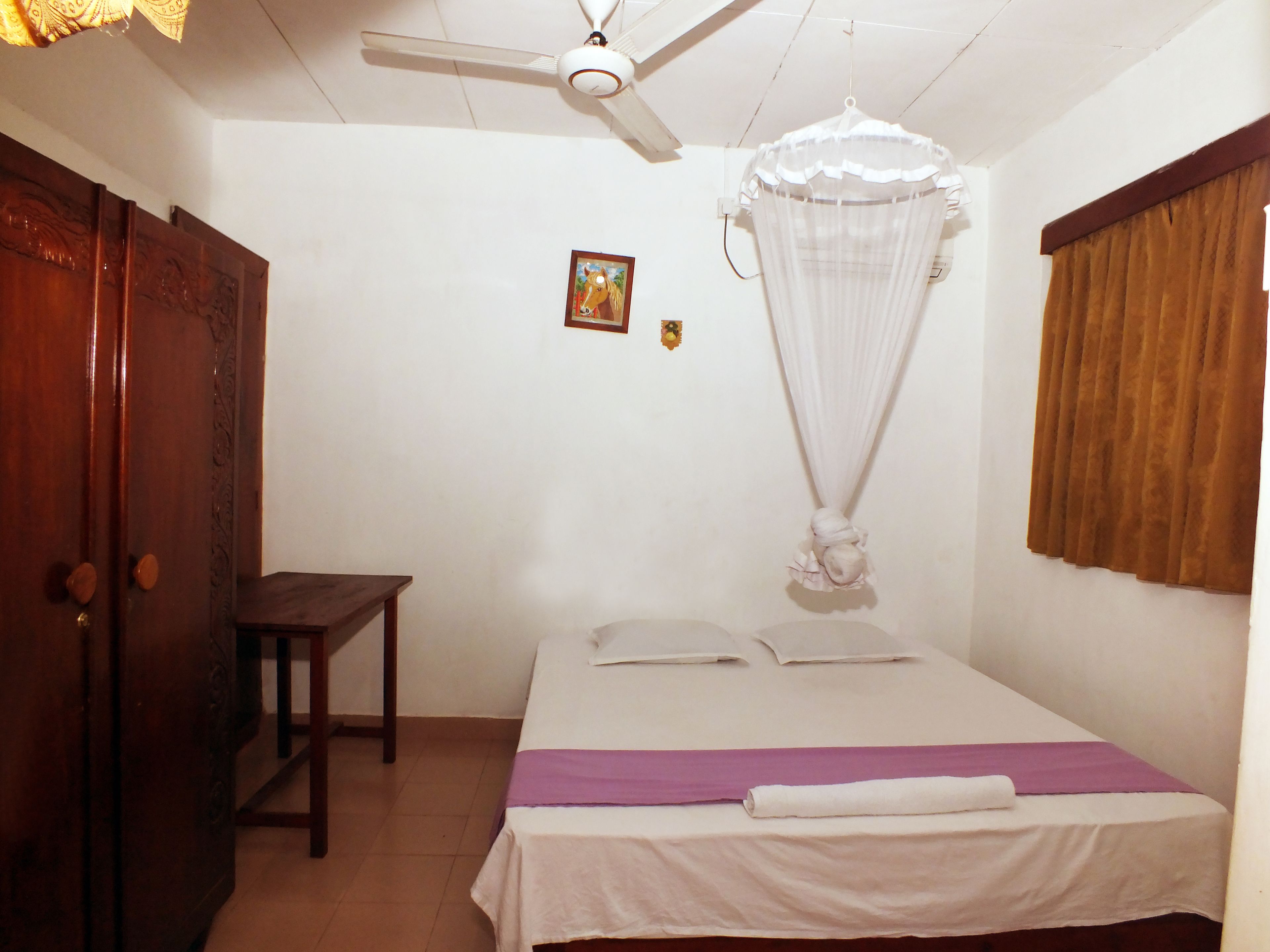 A bedroom with a bed, a ceiling fan, a wooden wardrobe, a small table, and a wall-mounted picture.