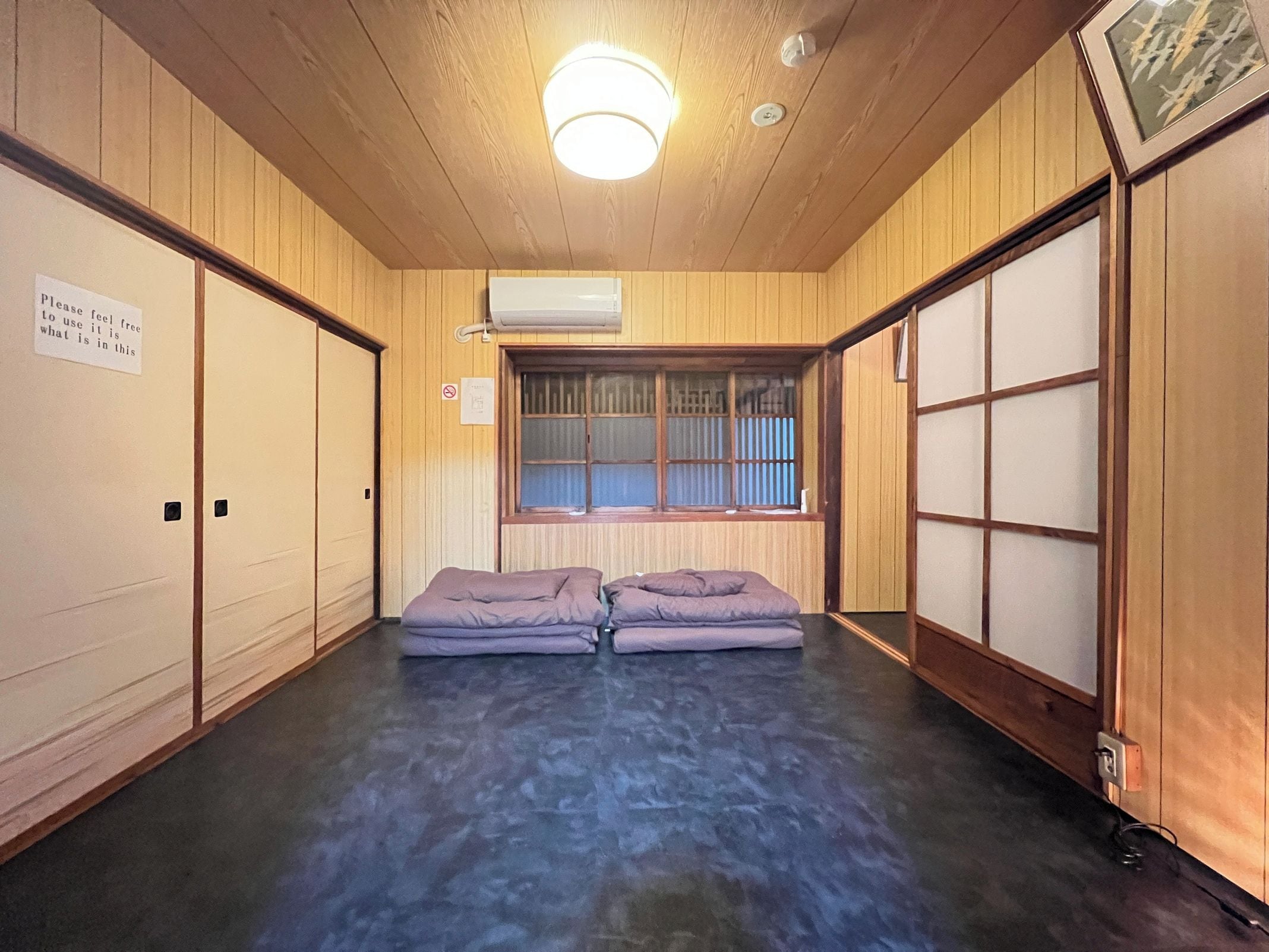 Ein Zimmer mit Holzwänden, einem Fenster mit Jalousien, einer Deckenleuchte und zwei Betten mit Kissen.