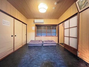 A room with wooden walls, a window with blinds, a ceiling light, and two beds with pillows.