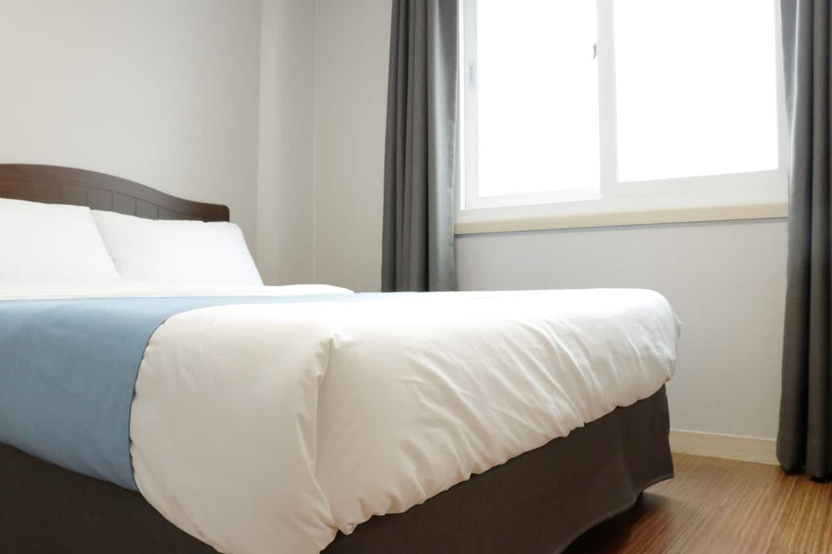 A neatly made bed with white and blue bedding, a wooden headboard, and a window with curtains.