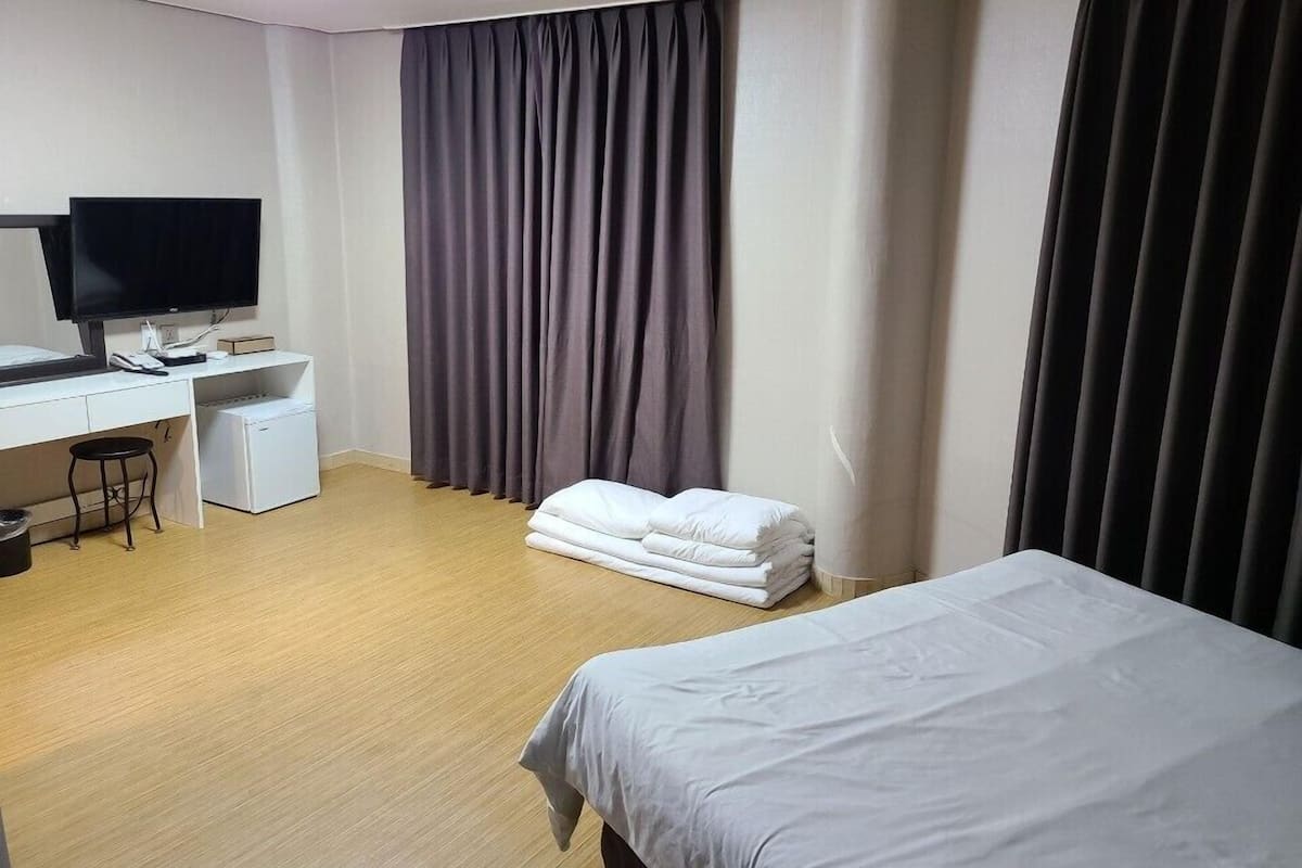 A hotel room with a bed, a desk with a computer, a chair, a small stool, a TV, and a window with curtains.