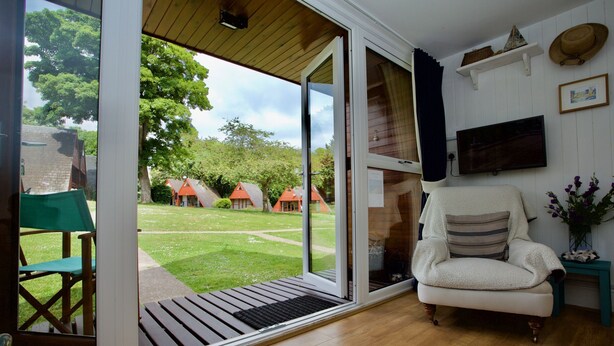 A room with a sliding glass door leading to a garden, a white armchair, a small table with a vase, and a mounted television. - Beach View - Kingsdown Resort (Kingsdown)