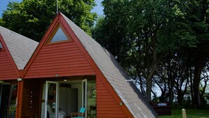 A red-roofed cabin with a glass door entrance, a small porch with a chair, and a wooden deck. - Beach View - Kingsdown Resort (Kingsdown)