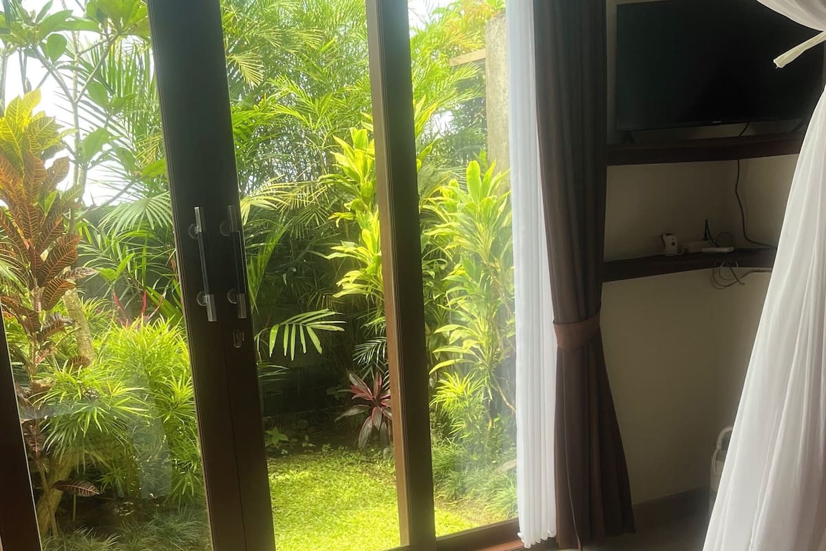 A hotel room with a bed, a view of a lush garden through a sliding glass door, and a television on a shelf.