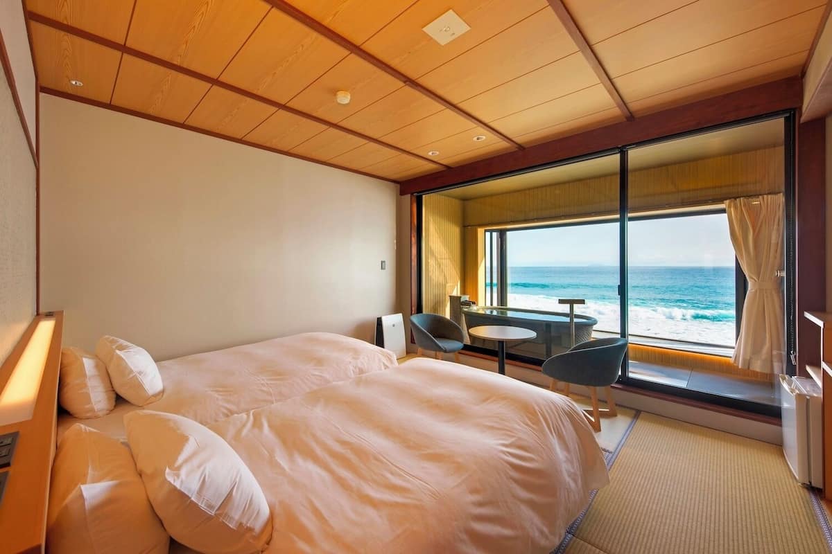 A hotel room with a bed, a small table, two chairs, a view of the ocean, and a large window.