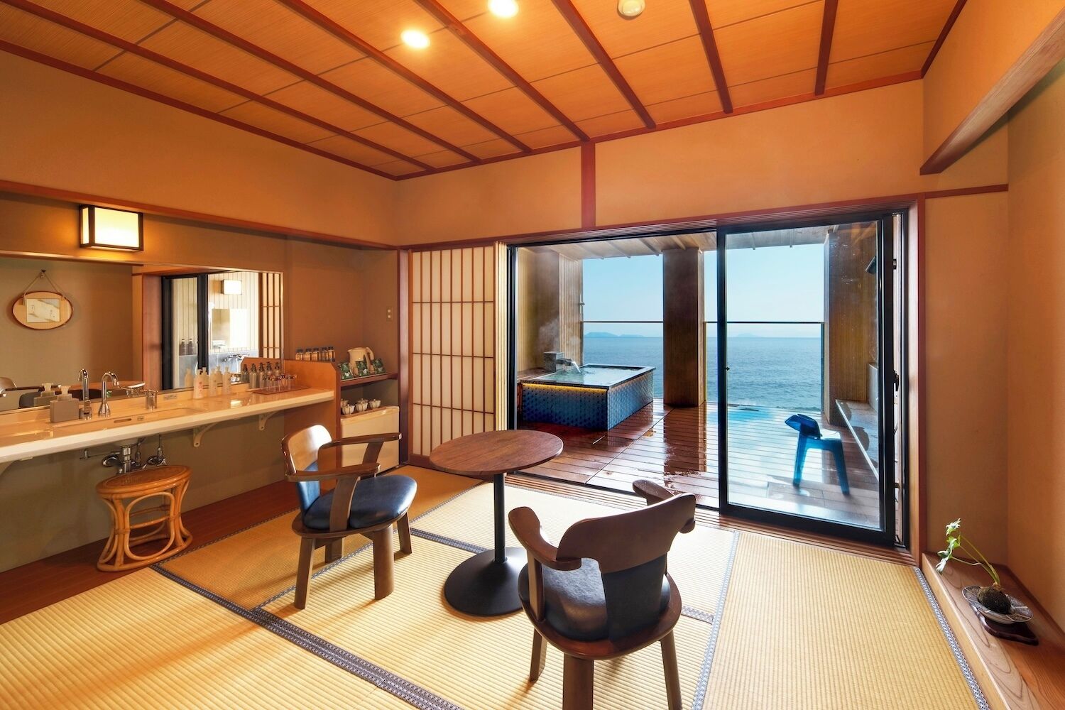 Habitación con techo de madera, un amplio ventanal corredero a un balcón, una pequeña mesa con sillas, un lavamanos con artículos de aseo y vista al mar.