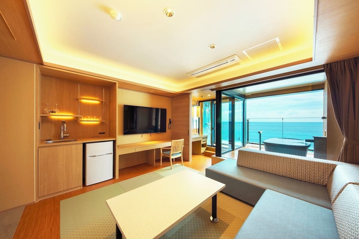 A modern hotel room with a dining area, a TV, a sofa, and a balcony with a view of the sea.