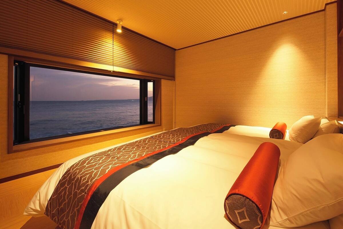 A bedroom with a large window, a bed with white bedding, and a view of the sea.