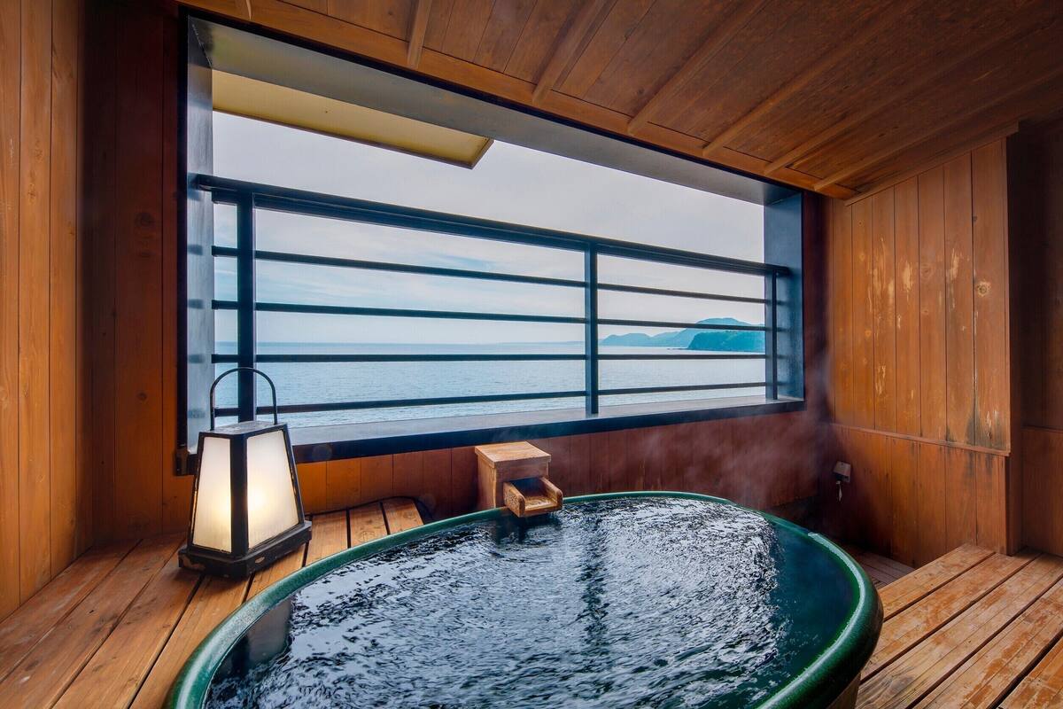 A wooden cabin with a hot tub, a lantern, and a view of the sea through a large window.
