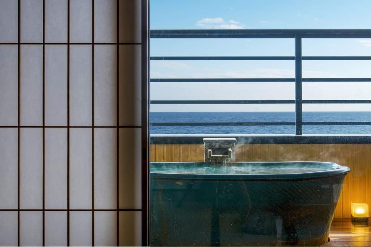 A bathtub with a view of the ocean through a window with horizontal blinds.
