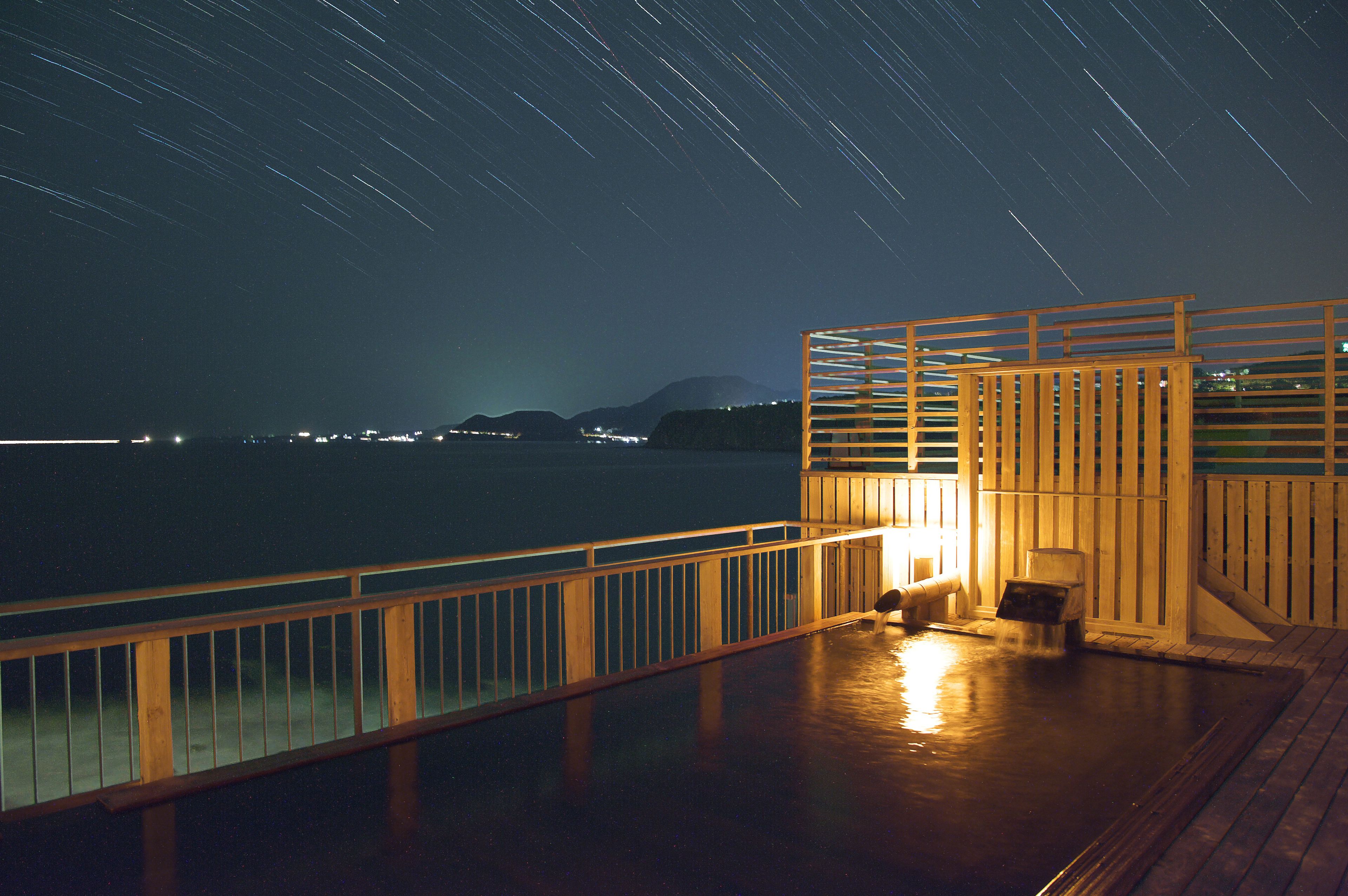 A wooden deck with a railing, a small seating area, and a view of the sea and mountains at night.