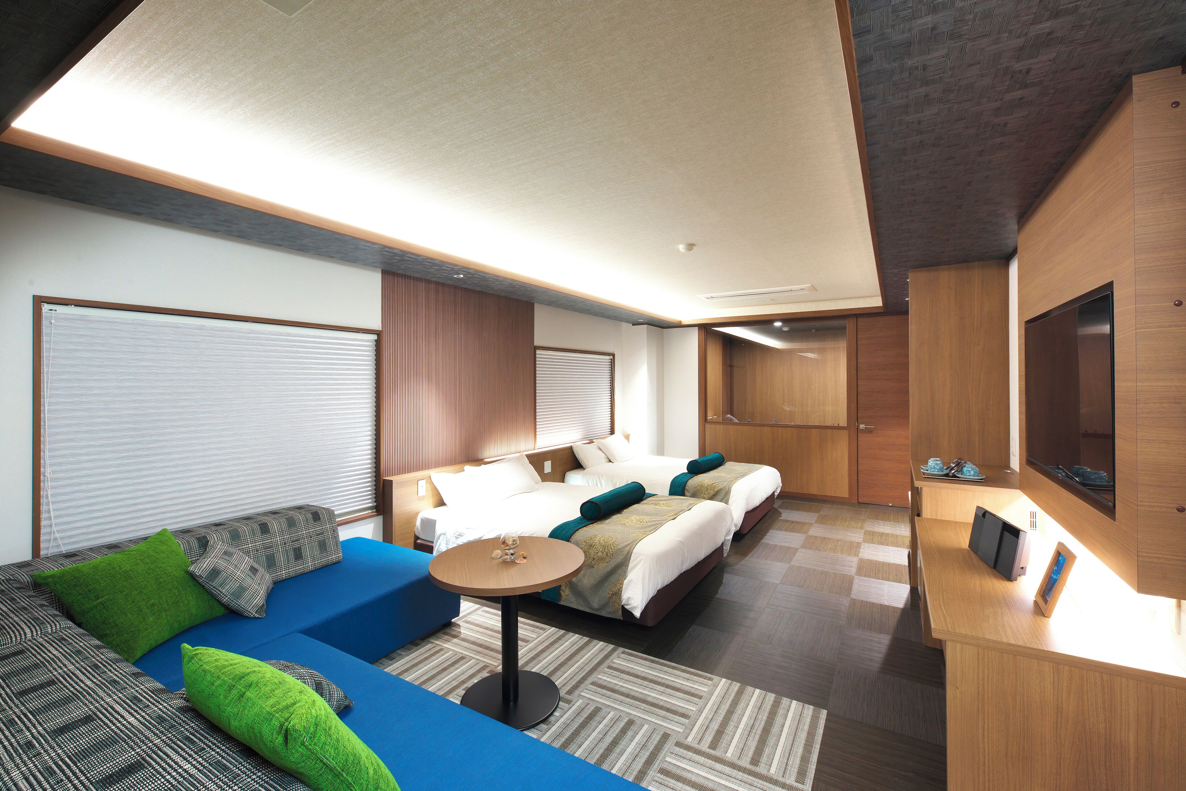 A modern hotel room with a sofa, two beds, a small table, and a desk with a flat-screen TV.