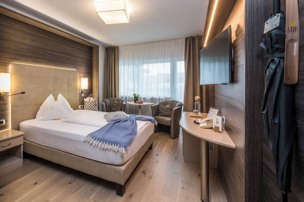 A modern hotel room with a large bed, bedside tables, a desk, a chair, a TV, and a wooden wall panel.