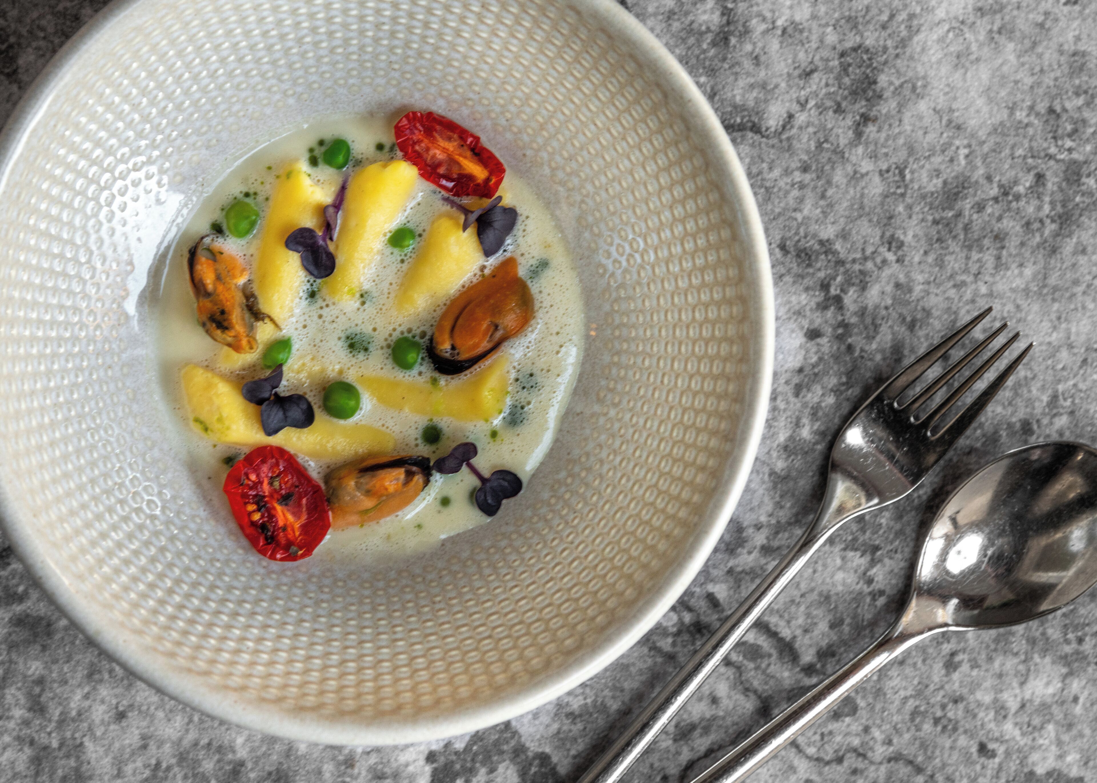 A bowl of soup with vegetables and mushrooms, accompanied by a fork and spoon on a marble surface.