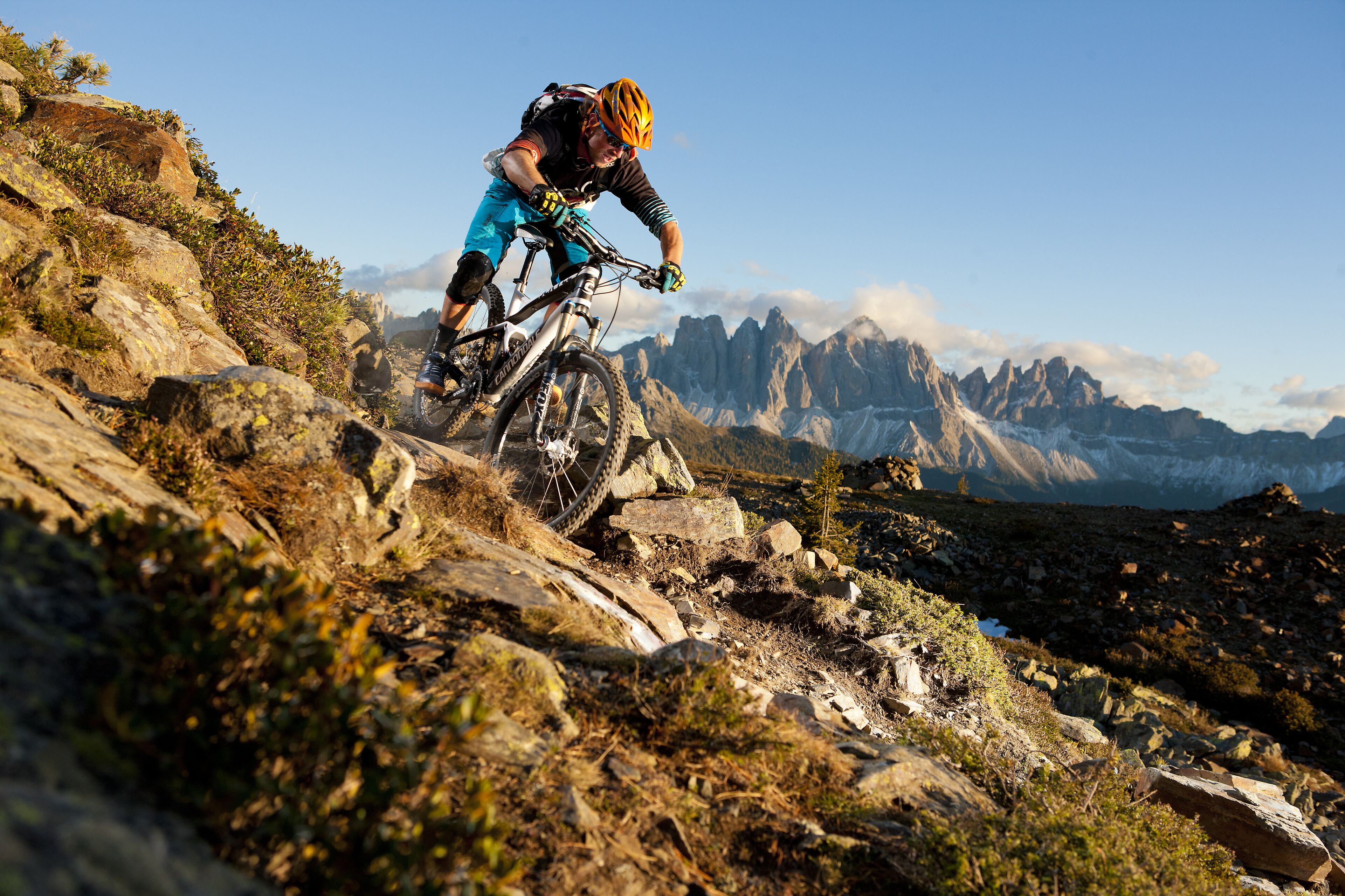 Un ciclista de montaña pedalea sobre un sendero rocoso, con montañas al fondo.