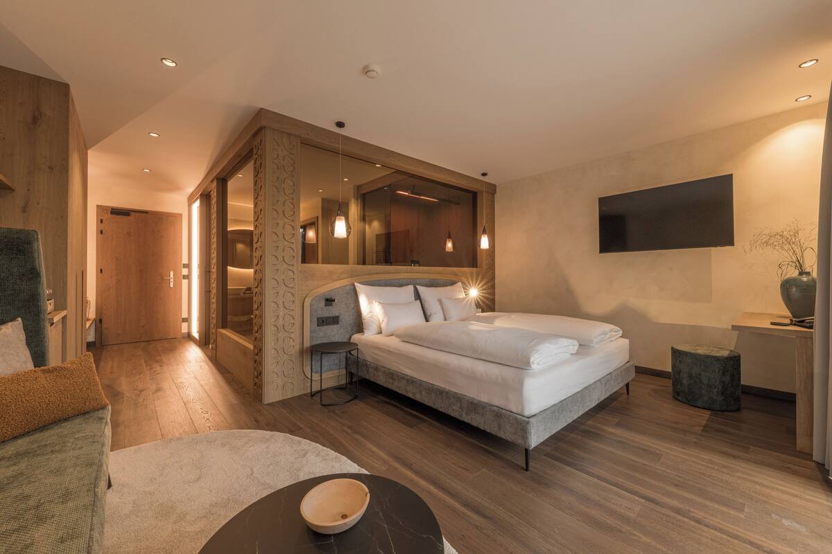 A modern hotel room with a bed, a seating area with a bowl, a TV mounted on the wall, and a wooden door leading to another room.