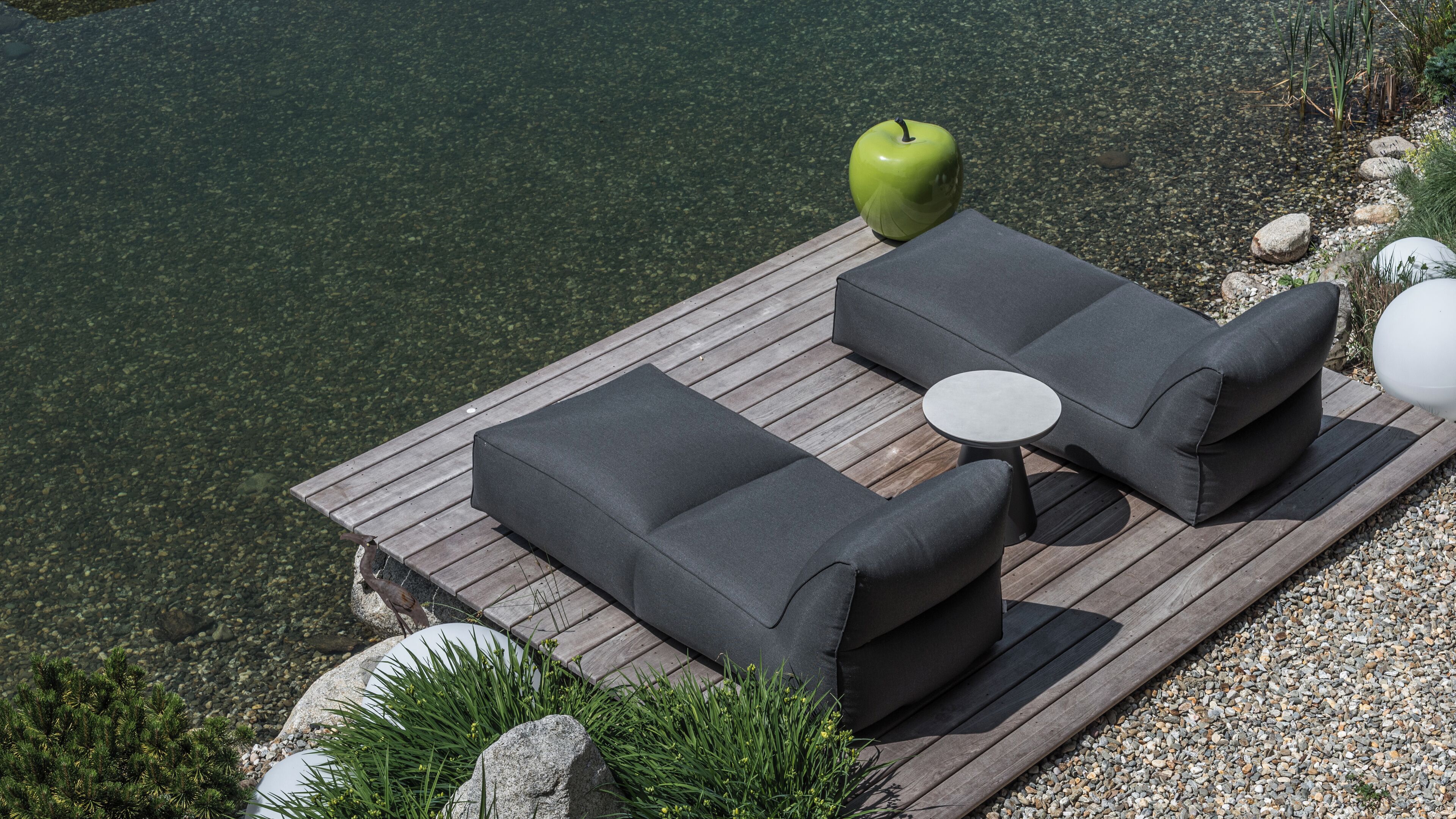 Eine Holzterrasse mit einer Couch und einem kleinen Tisch, mit Blick auf ein klares Gewässer.