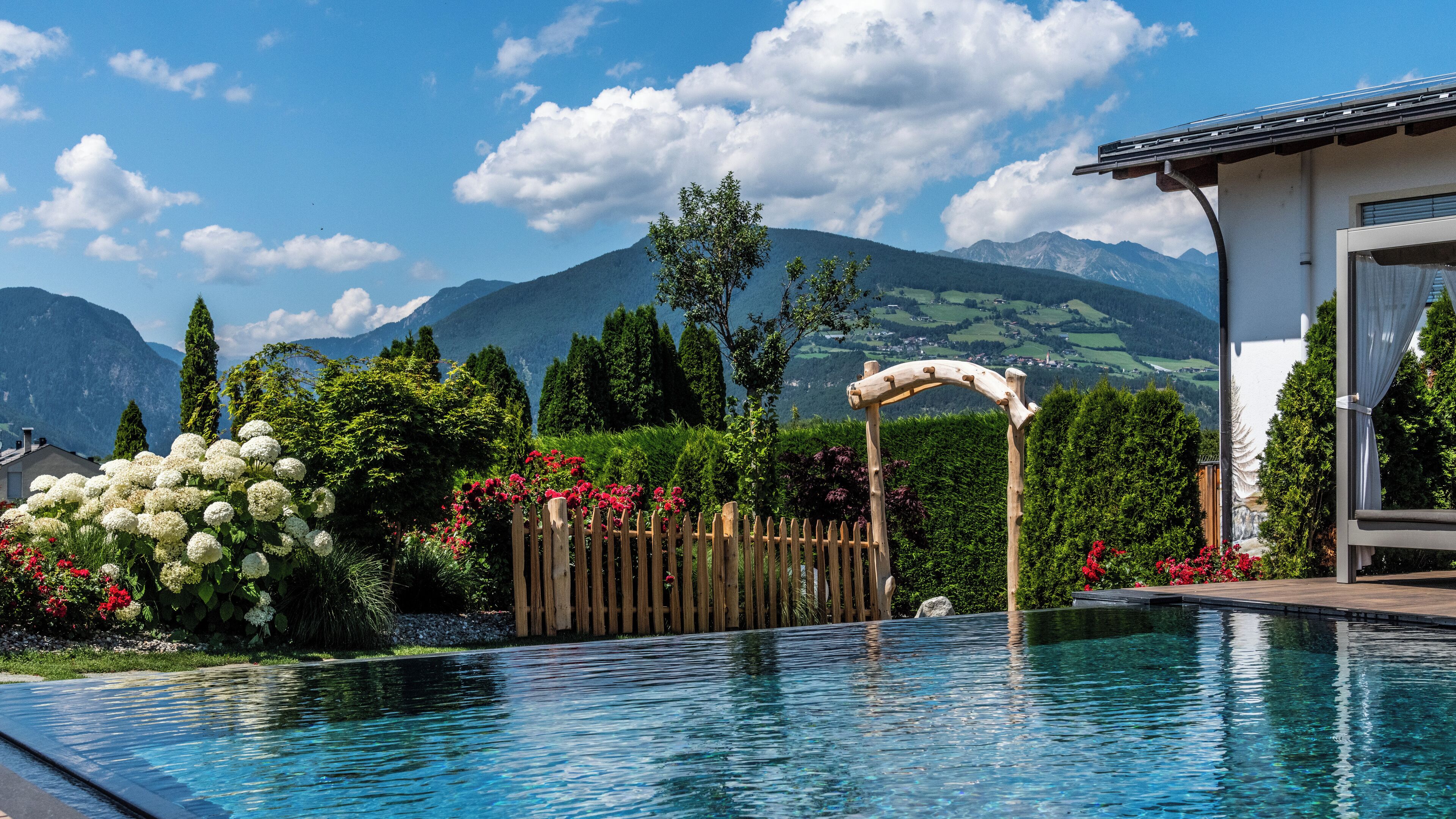 Ein Schwimmbecken mit Holzzaun und einem Bogen aus Holz, umgeben von üppigem Grün und Blumen, mit Bergen im Hintergrund.