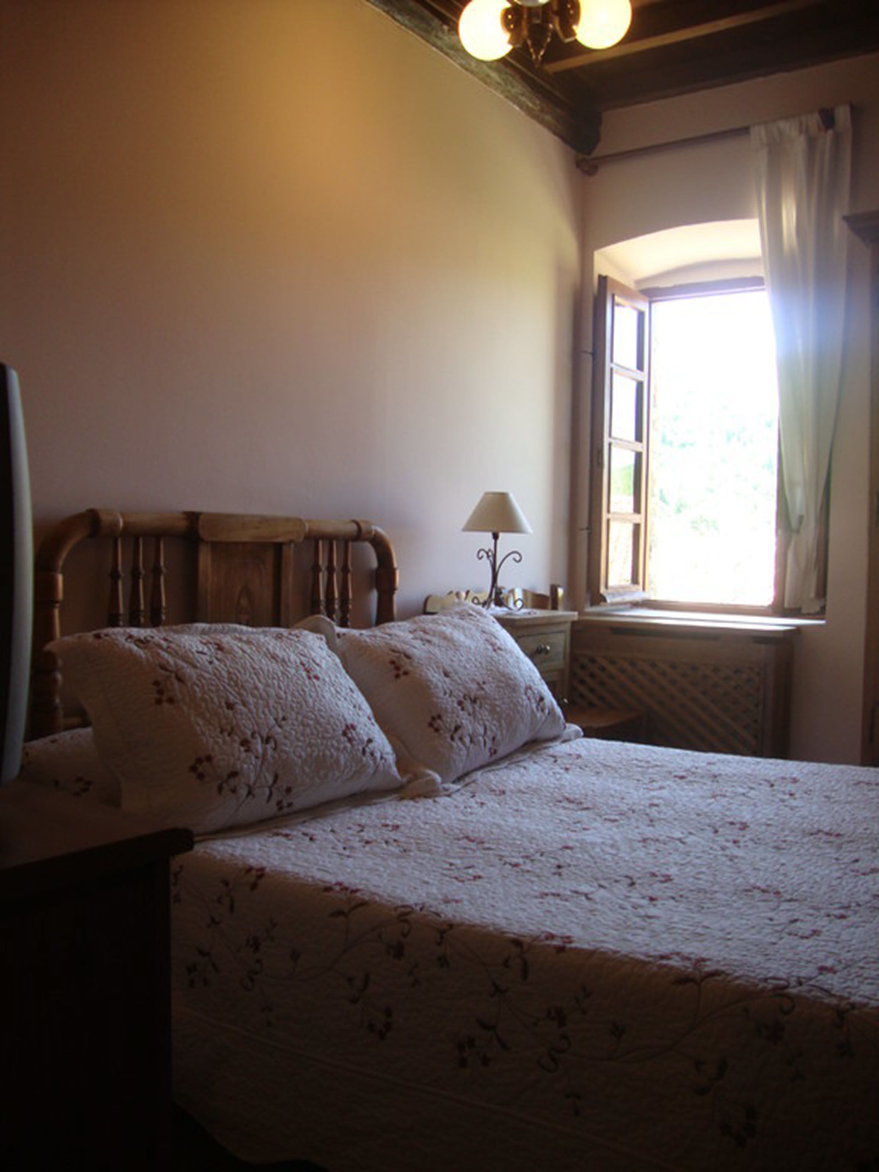Un dormitorio con cabecera de madera, cama con ropa de cama floral, mesita de noche con lámpara y ventana con cortinas.