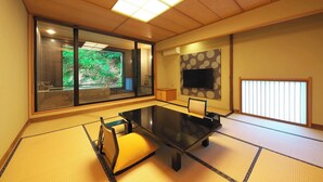 A traditional Japanese-style room with tatami flooring, a low table, a tatami mat chair, a television on the wall, and a sliding door leading to a bathroom with a view of greenery. - Kagiya the annex of Senoumi (Minamiizu)