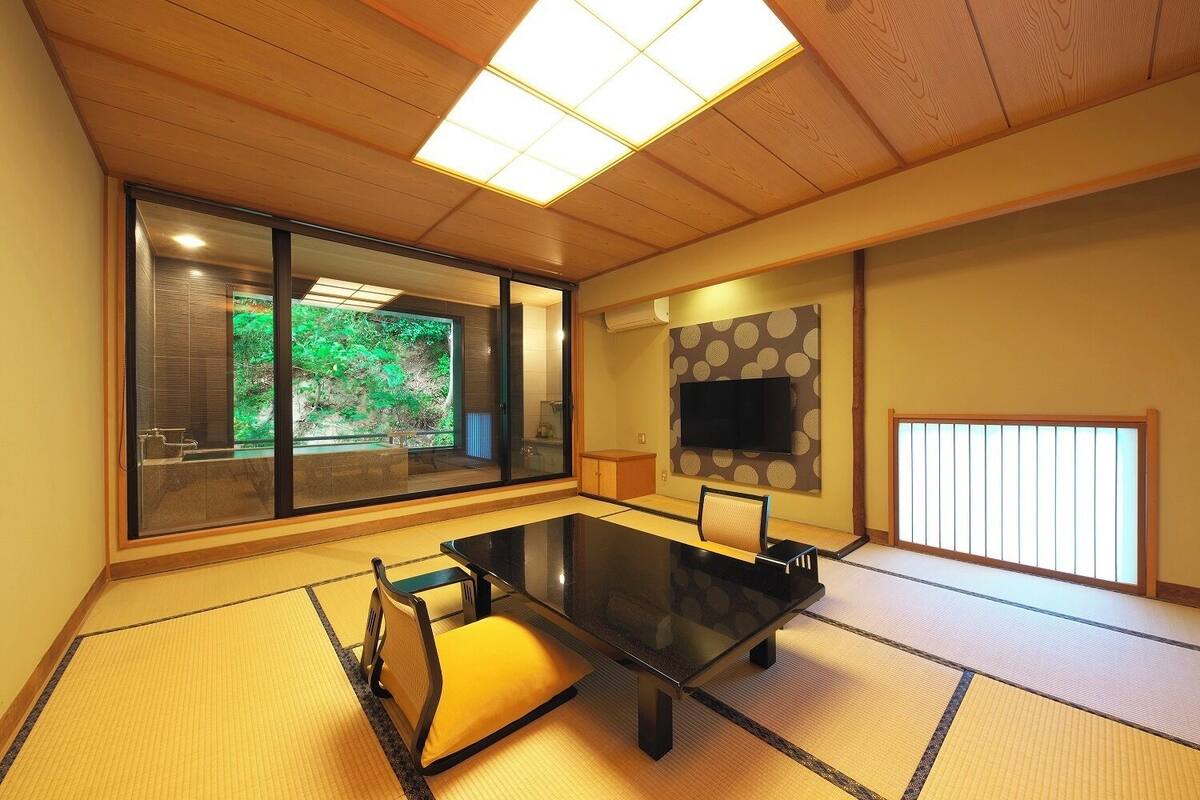 Ein traditioneller japanischer Raum mit Tatami-Bodenbelag, einem niedrigen Tisch, einem Tatami-Sitzkissen, einem an der Wand befestigten Fernseher und einer Schiebetür zu einem Badezimmer mit Blick auf Grünflächen.