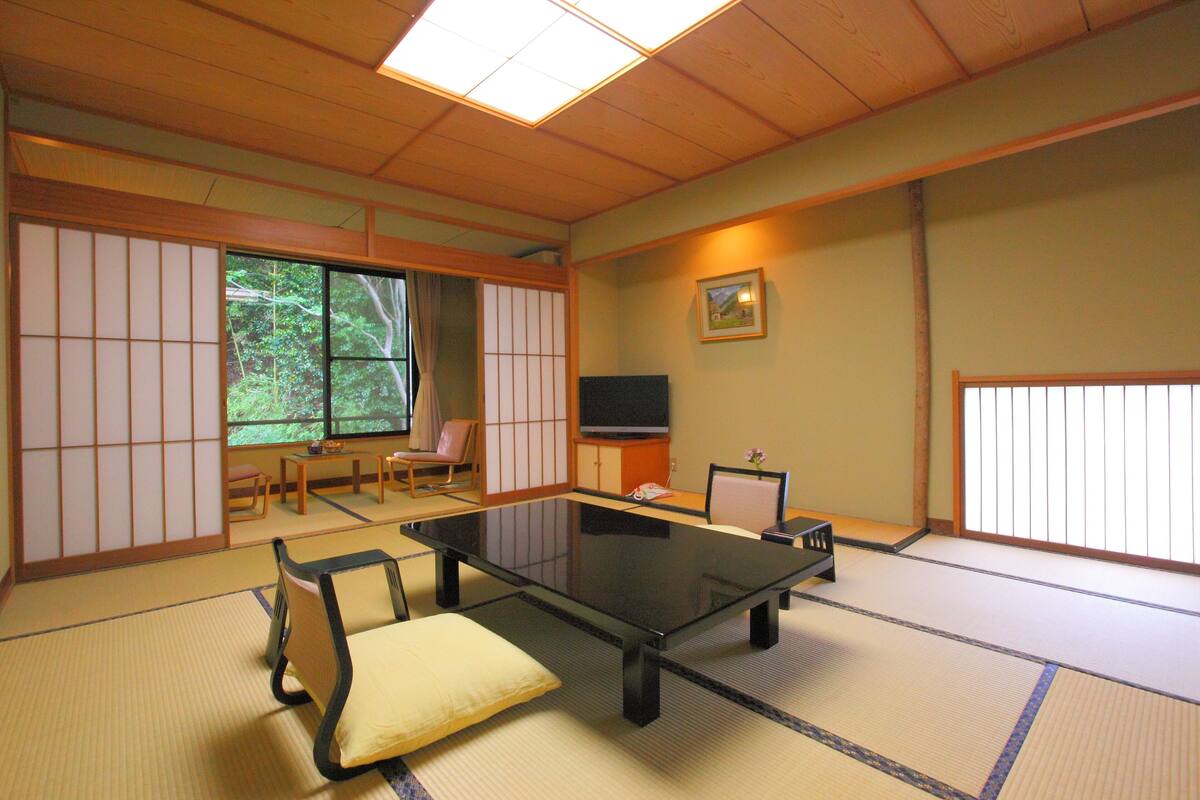 Ein Raum mit Tatami-Matten, einem niedrigen Tisch, Kissen, einem Fernseher und einer Schiebetür.