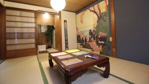 Una habitación japonesa tradicional con suelo de tatami, una mesa de madera baja con papeles, una lámpara colgante y una pintura grande en la pared.