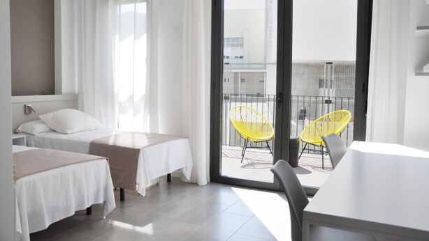 Habitación de hotel con dos camas, un balcón con sillas amarillas y vistas a edificios.