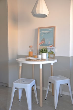 A small round table with a white tablecloth, two white stools, a glass bottle, a mug, a small potted plant, and a framed picture on the wall. - Residencia Universitaria Resa Tarragona Mediterrani - Campus Accomodation (Tarragona)