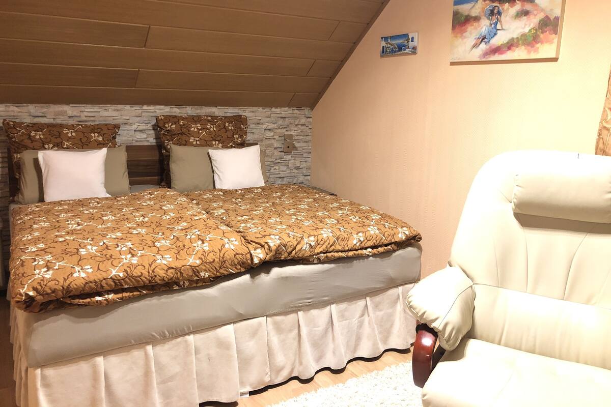 A bedroom with a wooden ceiling, a bed with a patterned bedspread, a white chair, and a painting on the wall.