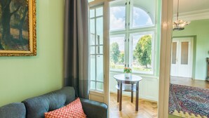 A living room with a sofa, a painting, a large window, and a view of the outdoors. - Riverside Apartment With A Balcony (Prague)