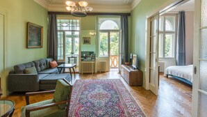 A spacious living room with a large sofa, a coffee table, and a patterned rug. There is a television on a wooden cabinet, and a door leading to another room. - Riverside Apartment With A Balcony (Prague)