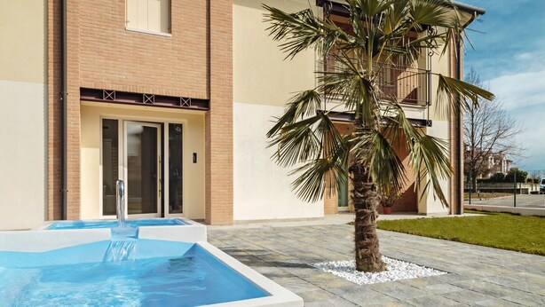 A swimming pool with a palm tree in the foreground, a brick building with a glass door entrance, and a paved patio area. - Cimadolmo Prosecco and History (Cimadolmo)