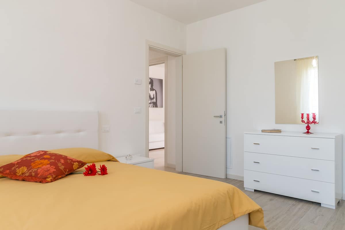 Quarto com cama, mesa de cabeceira, cômoda e porta que dá para outro espaço.
