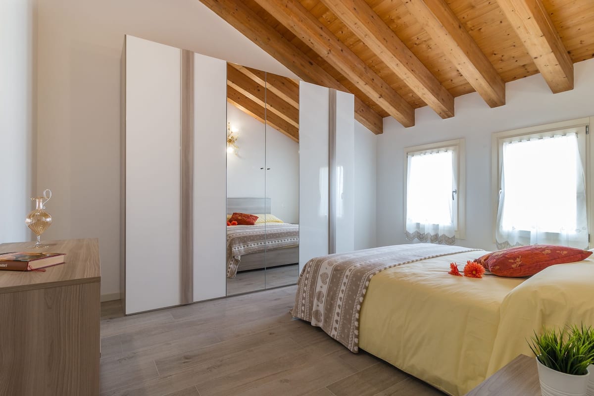 Quarto com teto de madeira, cama com colcha amarela, mesa de cabeceira com um livro e janela com cortinas.