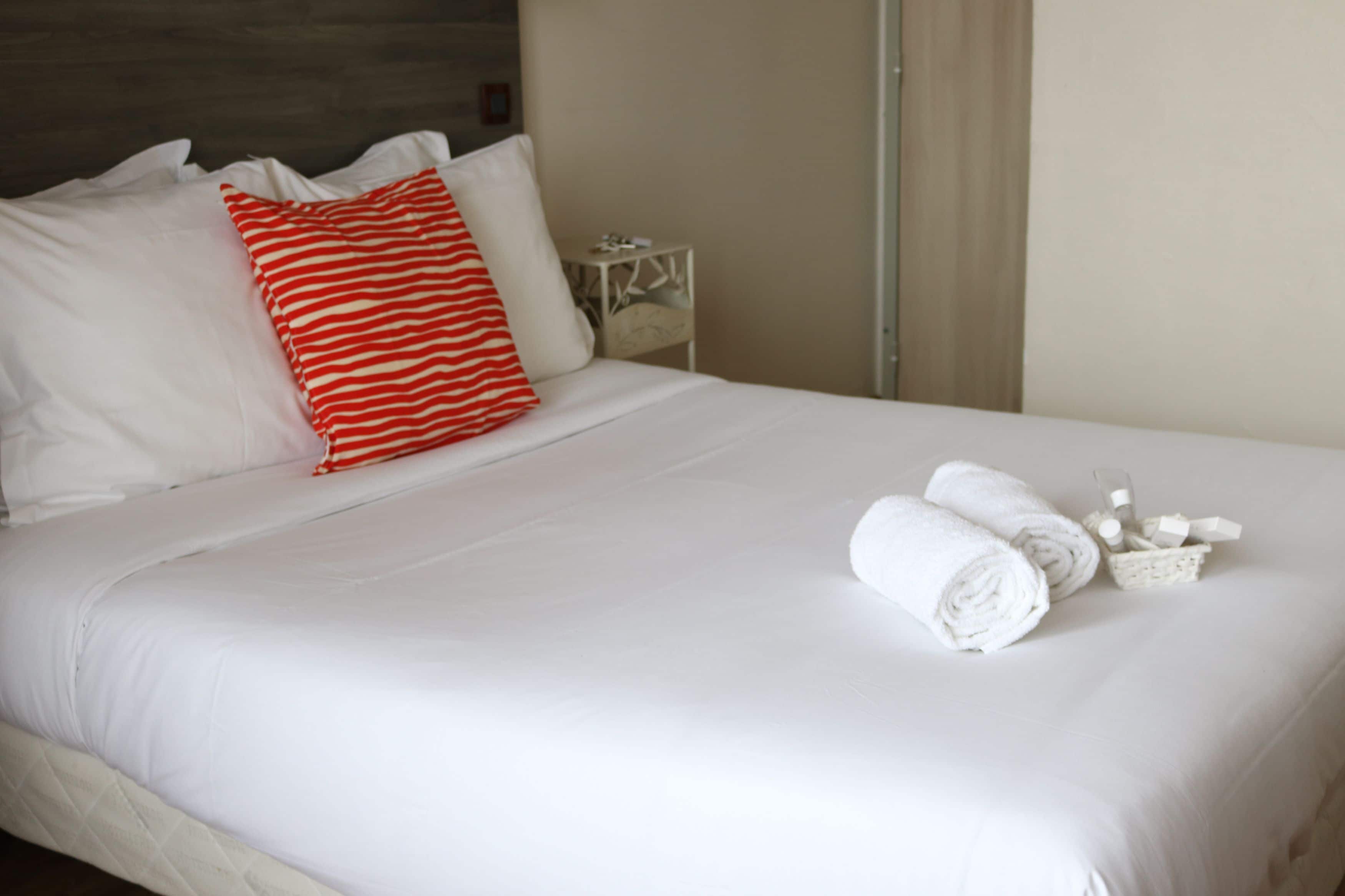 A neatly made bed with white bedding and a red and white striped pillow, a small bedside table with a lamp, and rolled towels on the bed.