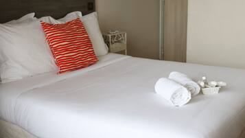 A neatly made bed with white bedding and a red and white striped pillow, a small bedside table with a lamp, and rolled towels on the bed.