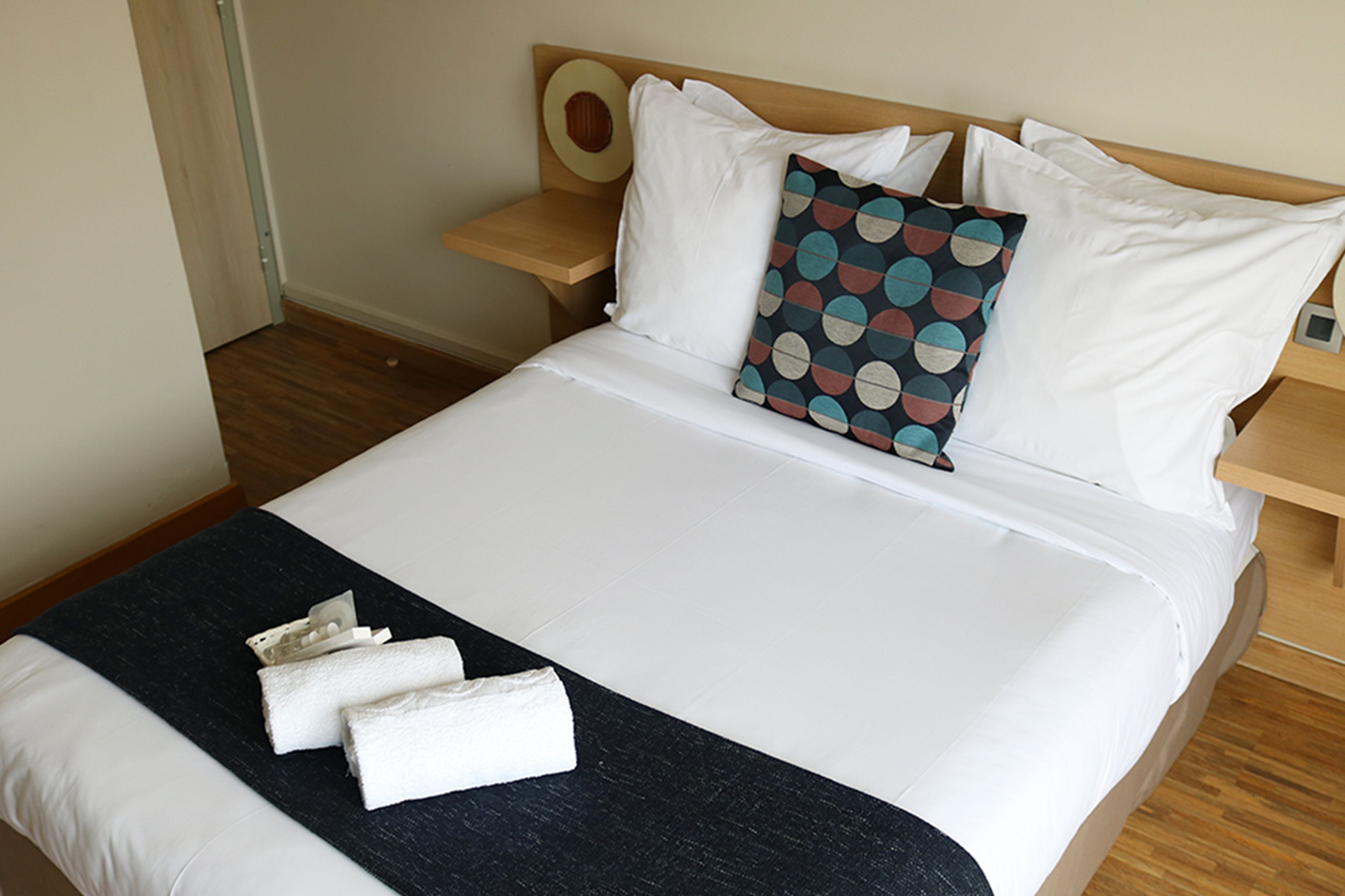 A neatly made bed with white linens, a black throw blanket, and a pillow with a colorful geometric pattern.