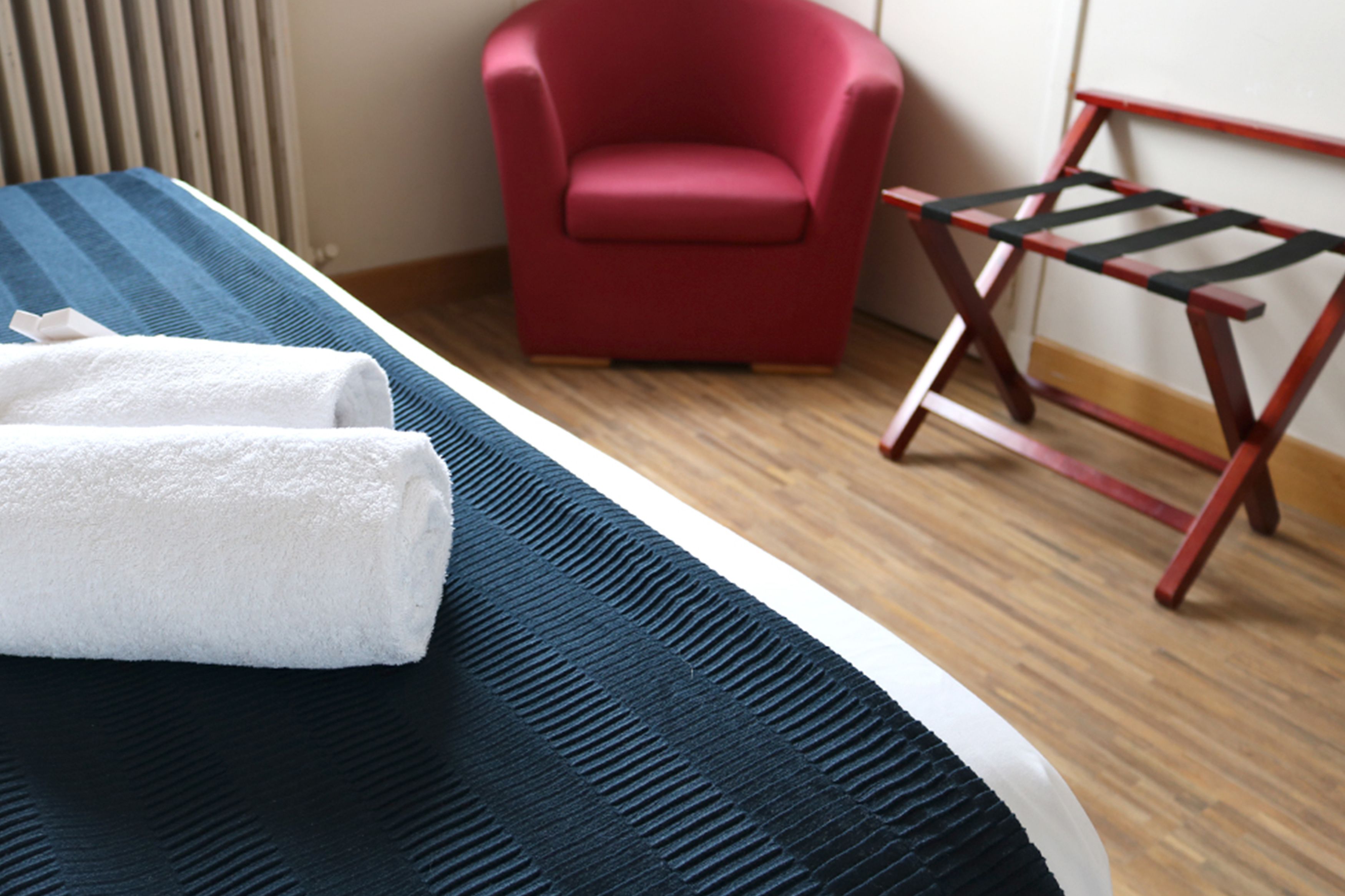 A neatly made bed with a blue and black striped bedspread, a white towel, and a red chair in a room with wooden flooring and a radiator.