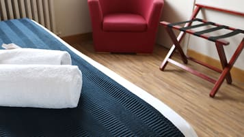 Ein ordentlich bezogenes Bett mit einer blau-schwarz gestreiften Tagesdecke, ein weißes Handtuch und ein roter Sessel in einem Zimmer mit Holzboden und Heizkörper.