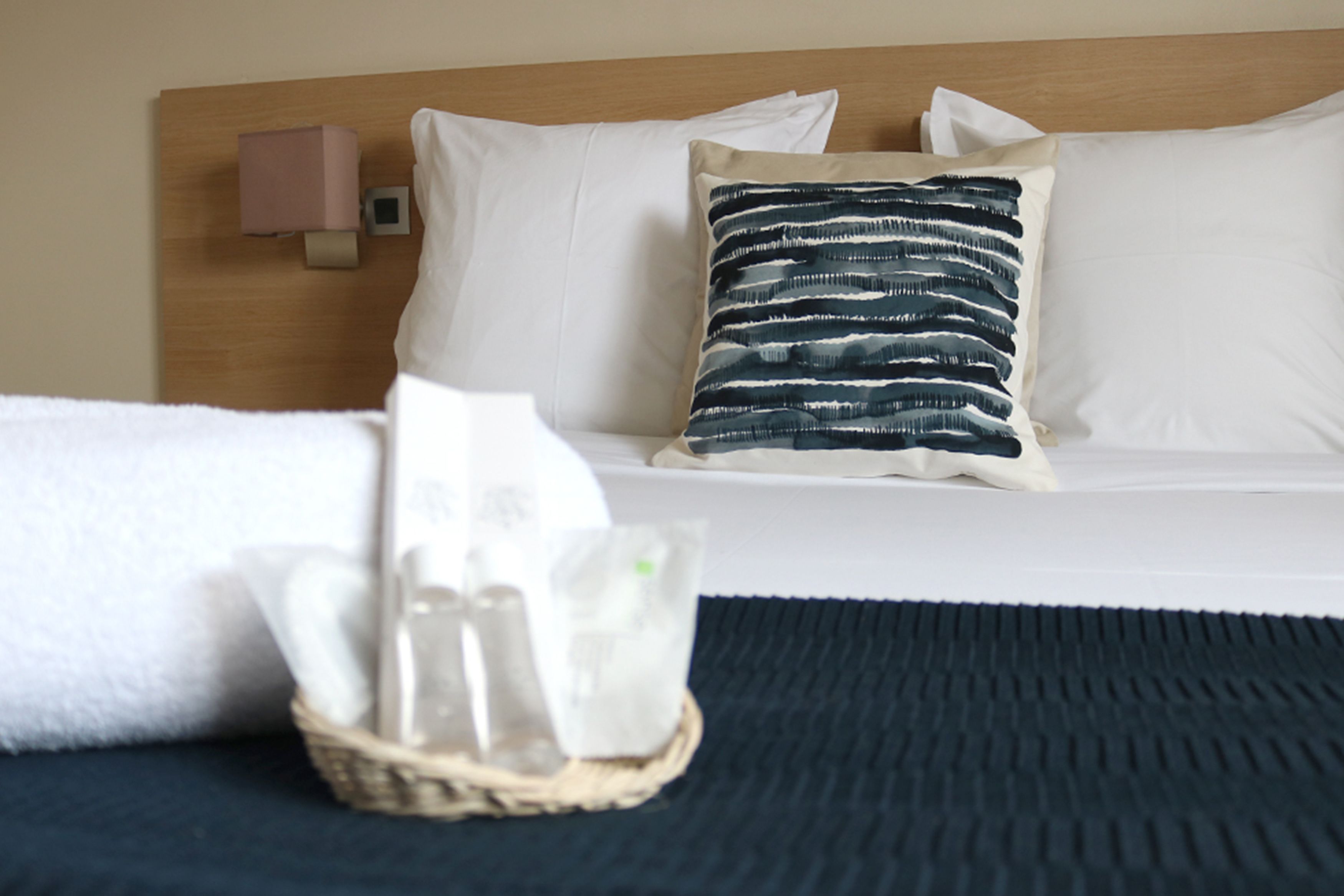 A neatly made bed with white and striped pillows, a bedside table with a glass and a tissue box, and a wooden headboard.