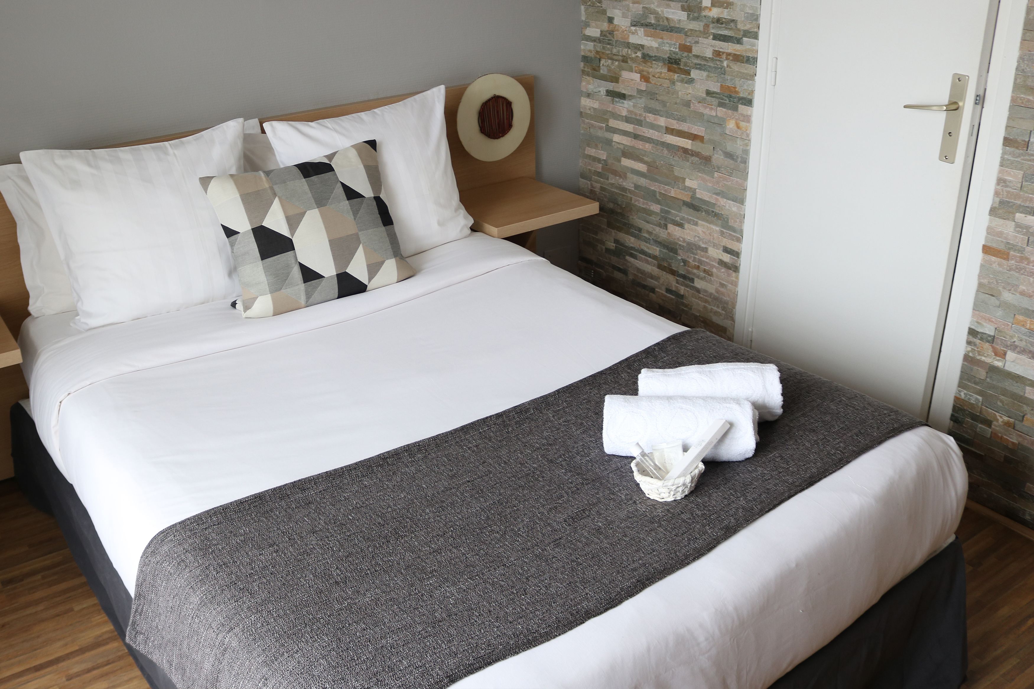A neatly made bed with white and patterned pillows, a grey throw blanket, and a folded towel on the foot of the bed.