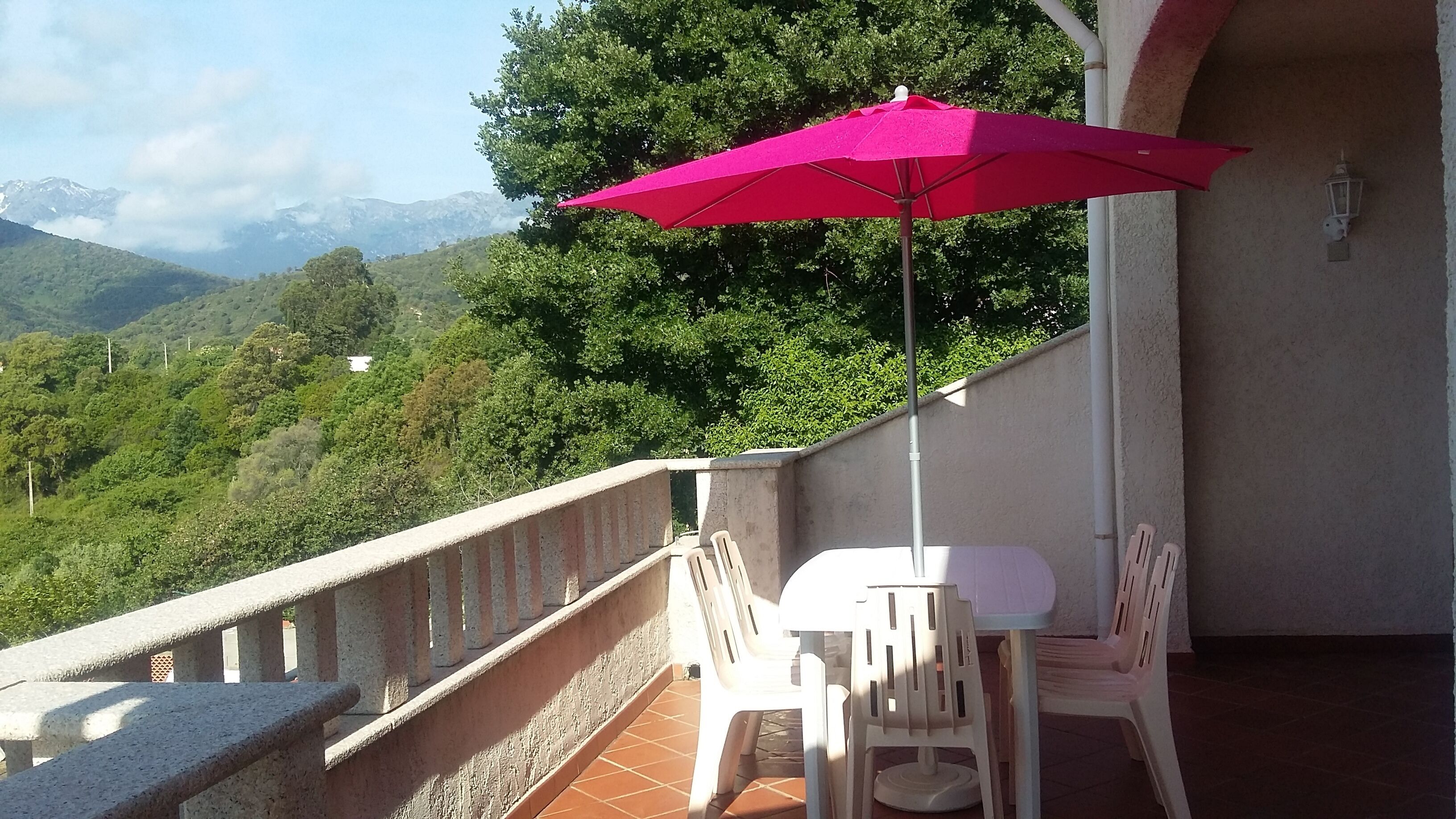 En balkon med rød parasol, hvide stole og udsigt til grønne bakker og bjerge.