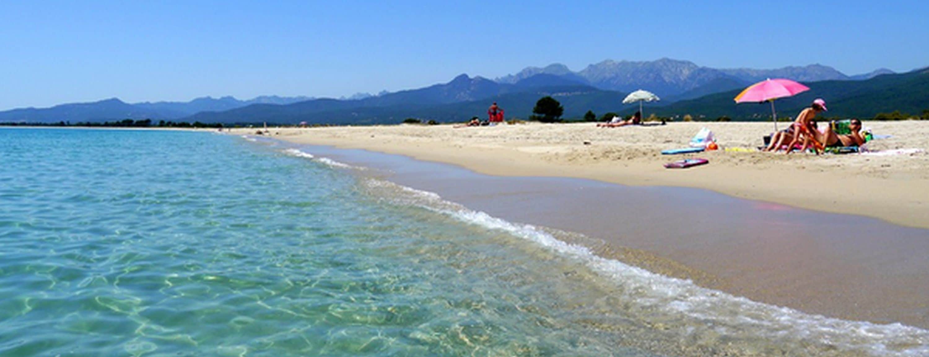 A beach with clear water, a sandy shore, and mountains in the background.