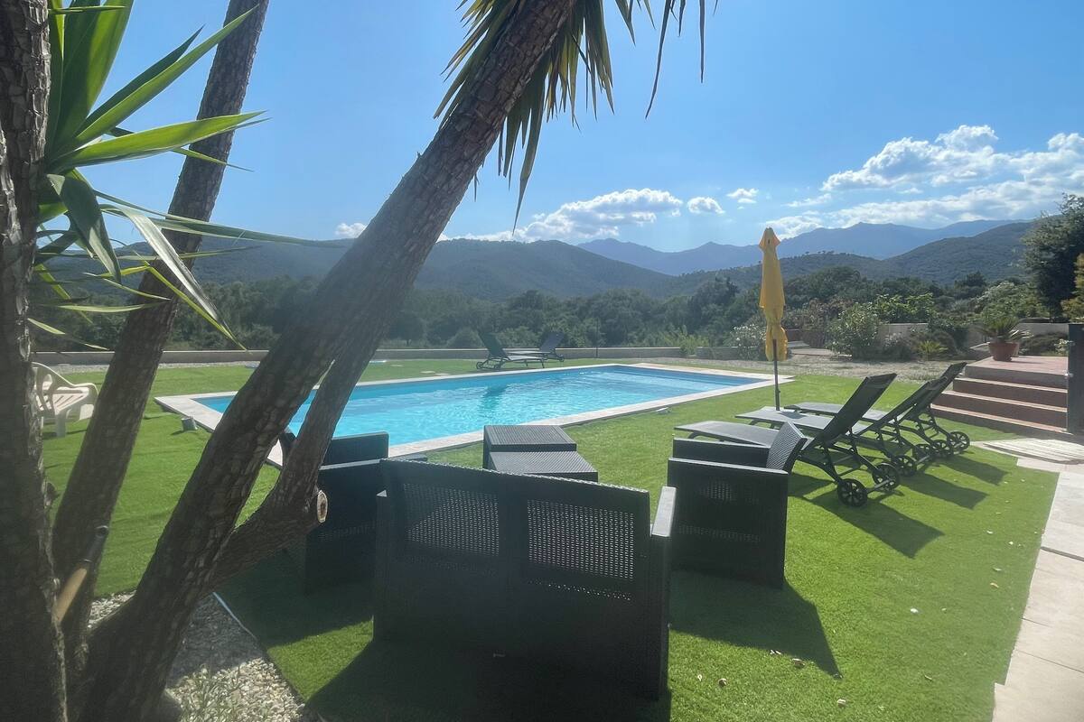 Un área de piscina con vistas a montañas, césped verde y sillones de descanso.